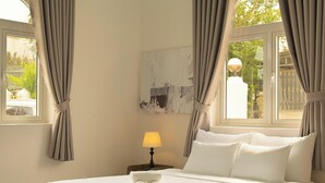 Quarto de hotel com cama, mesa de cabeceira com luminária, janela com cortinas e vista para o verde lá fora.