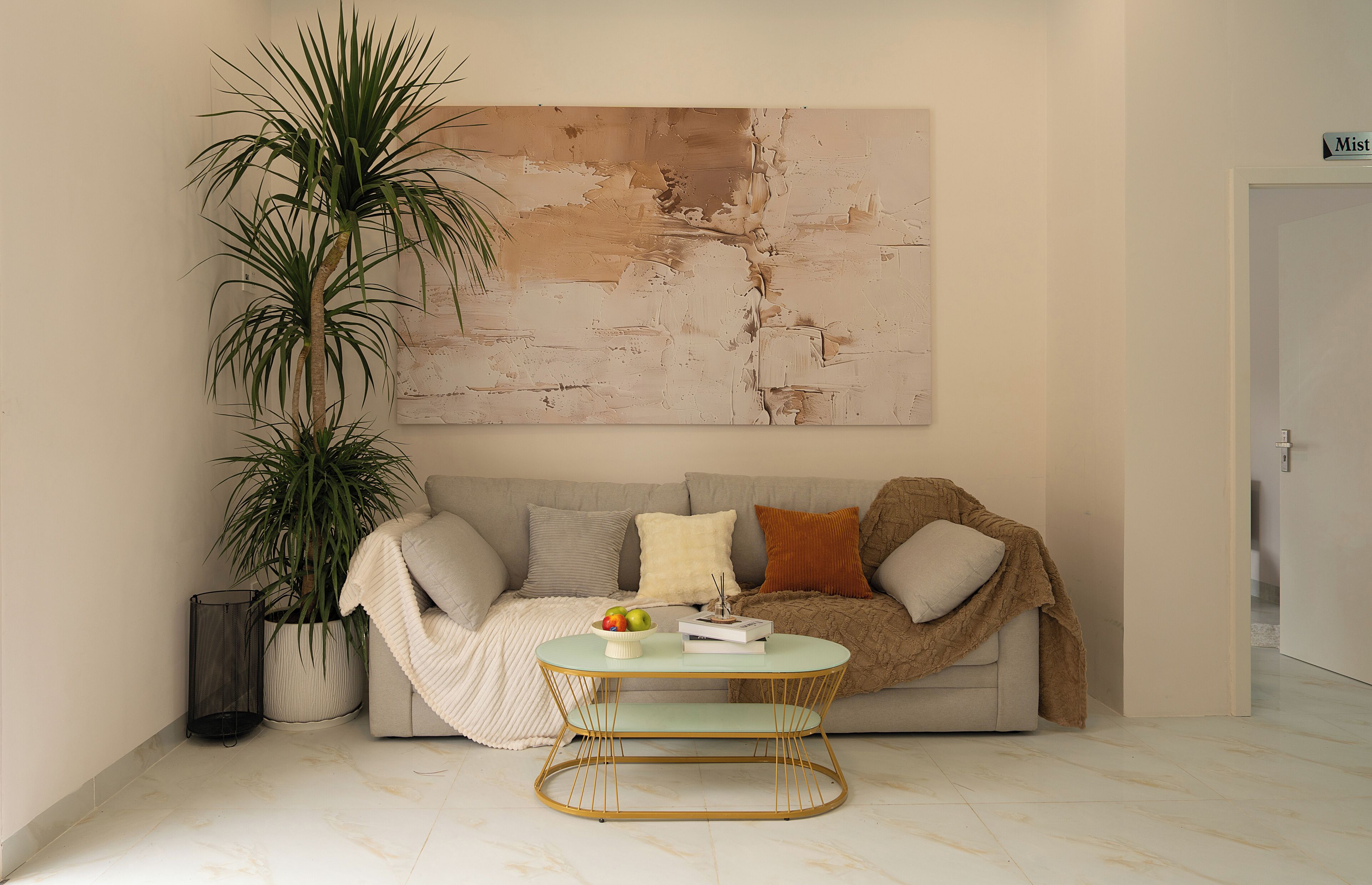 Ein Wohnzimmer mit einer Couch, einem Couchtisch, einer großen Pflanze und einem Wandkunstwerk mit Struktur.