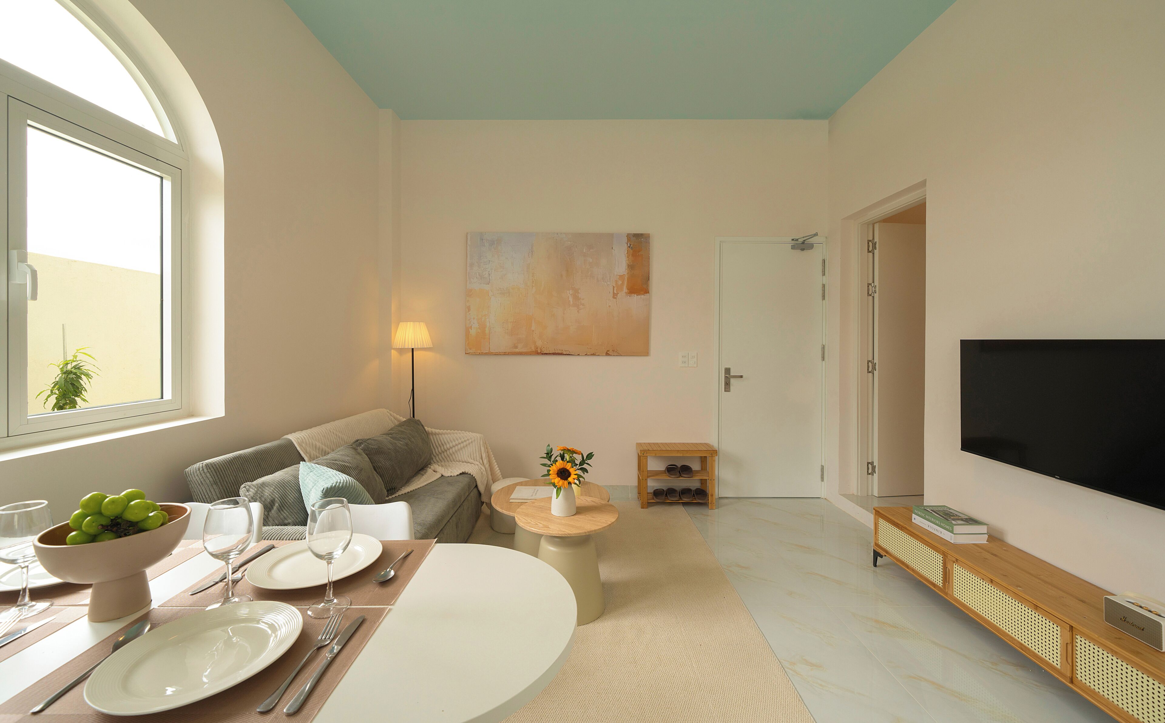 Ein Wohnzimmer mit einer Couch, einem Couchtisch, einem Esstisch gedeckt mit Tellern und Gläsern, einer Lampe, einem Bild an der Wand und einem Fernseher an der Wand.