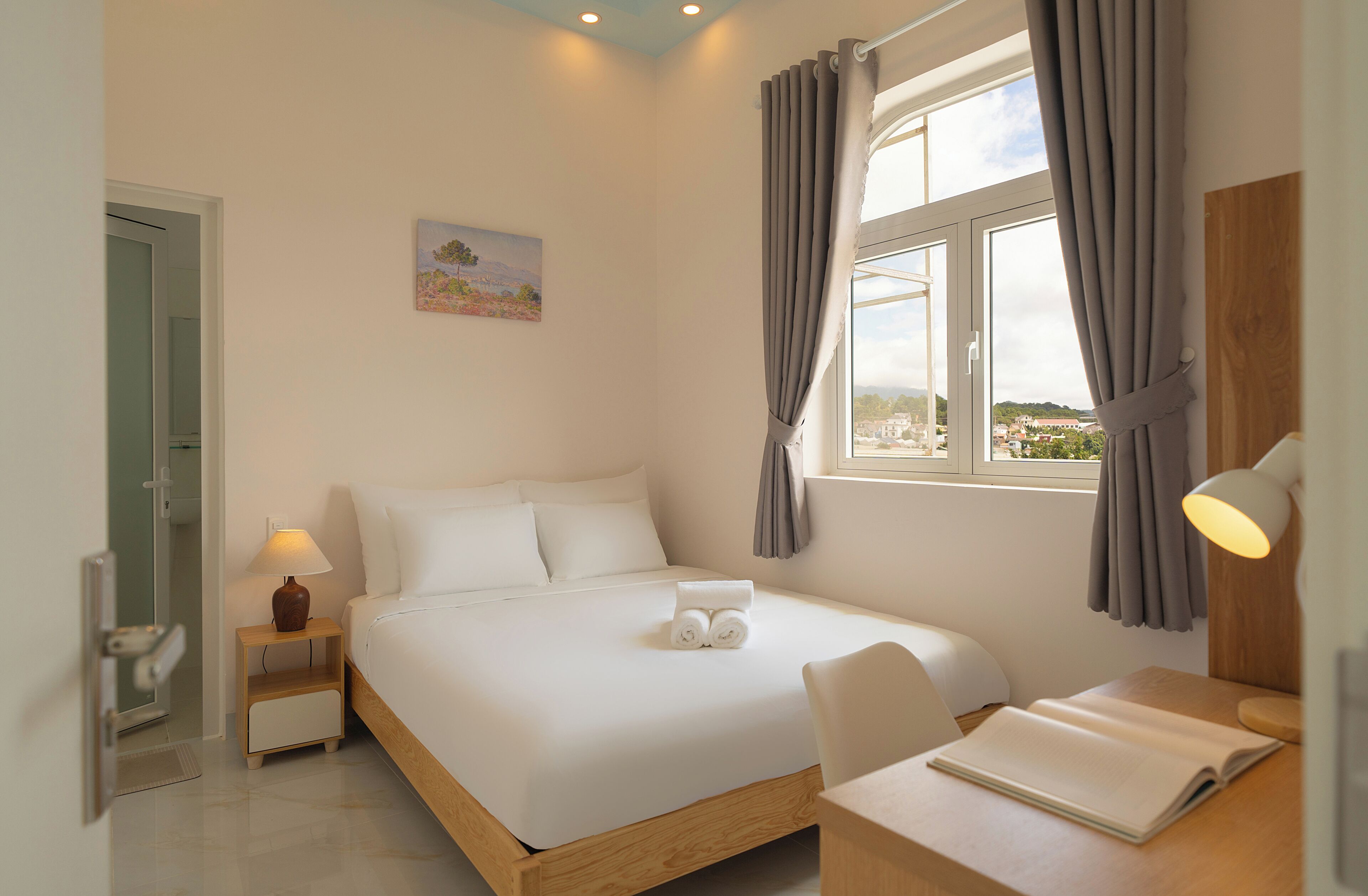 A hotel room with a bed, a desk, a chair, a window with curtains, a bedside lamp, and a painting on the wall.