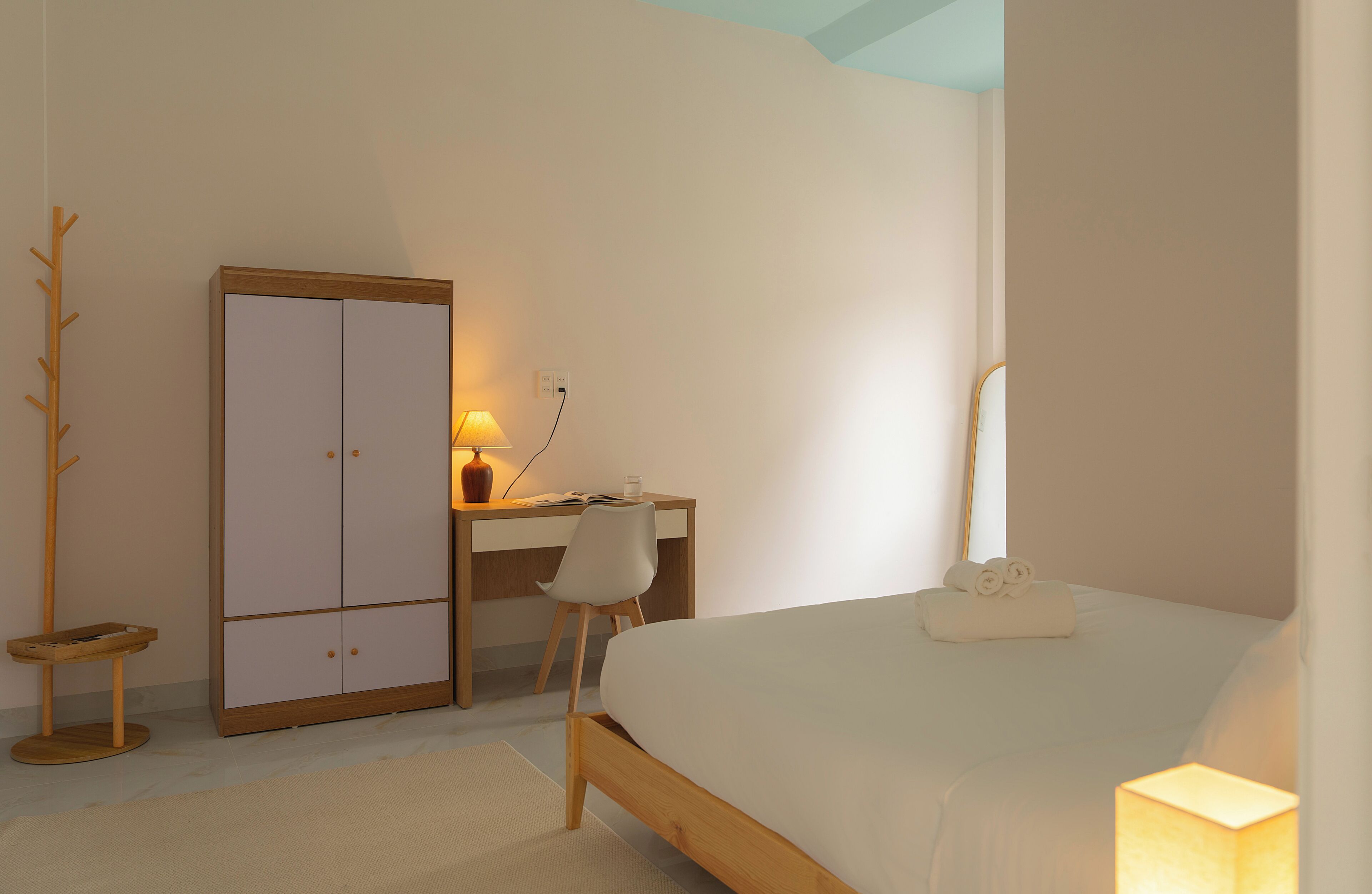 A bedroom with a bed, a desk with a chair, a wardrobe, and a bedside table with a lamp.