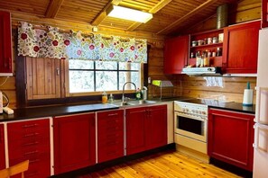A kitchen with wooden cabinets, a stove, and a window with a floral curtain. - Log cabin in a secluded location (Ulricehamn)