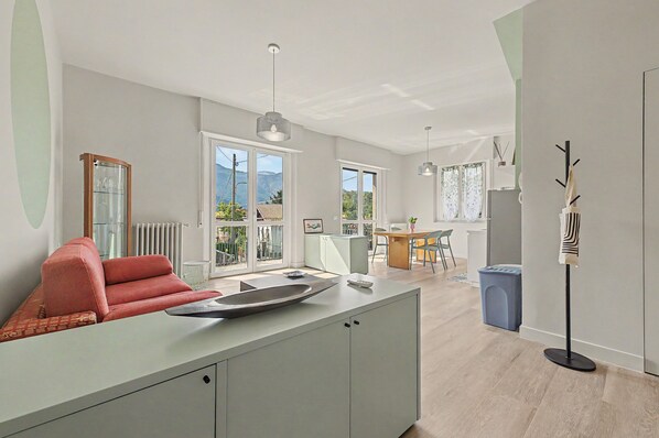 A modern living room with a sofa, a dining area with a table and chairs, and a view of the outdoors through large windows.