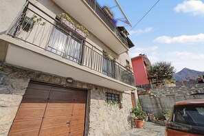 Edificio de dos pisos con fachada de piedra, puerta de garaje de madera y un balcón con plantas en macetas.