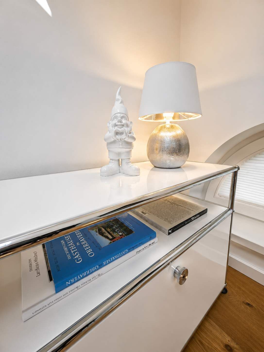 Ein weißer Nachttisch mit einer Lampe, einer dekorativen Zwergfigur und einem Buch mit dem Titel 'Gasthof BRABERER'.