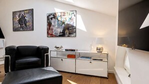 A modern living room with a skylight, a black leather sofa, a glass coffee table, and two wall-mounted paintings.