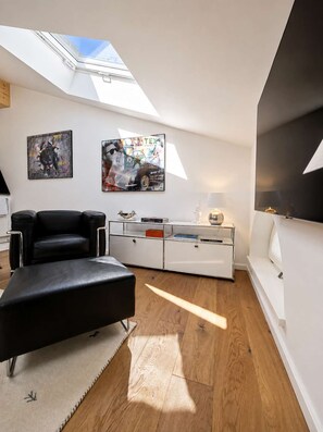 A modern living room with a skylight, a black leather sofa, a glass coffee table, and two wall-mounted paintings.