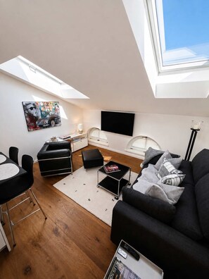 A modern living room with a skylight, a flat-screen TV, a black sofa, a dining area with a table and chairs, and a small side table with a lamp.