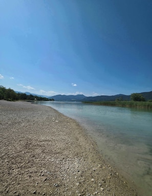 Une rive de rivière bordée d’eau claire, de galets et, au loin, de montagnes se dressant sous un ciel bleu.