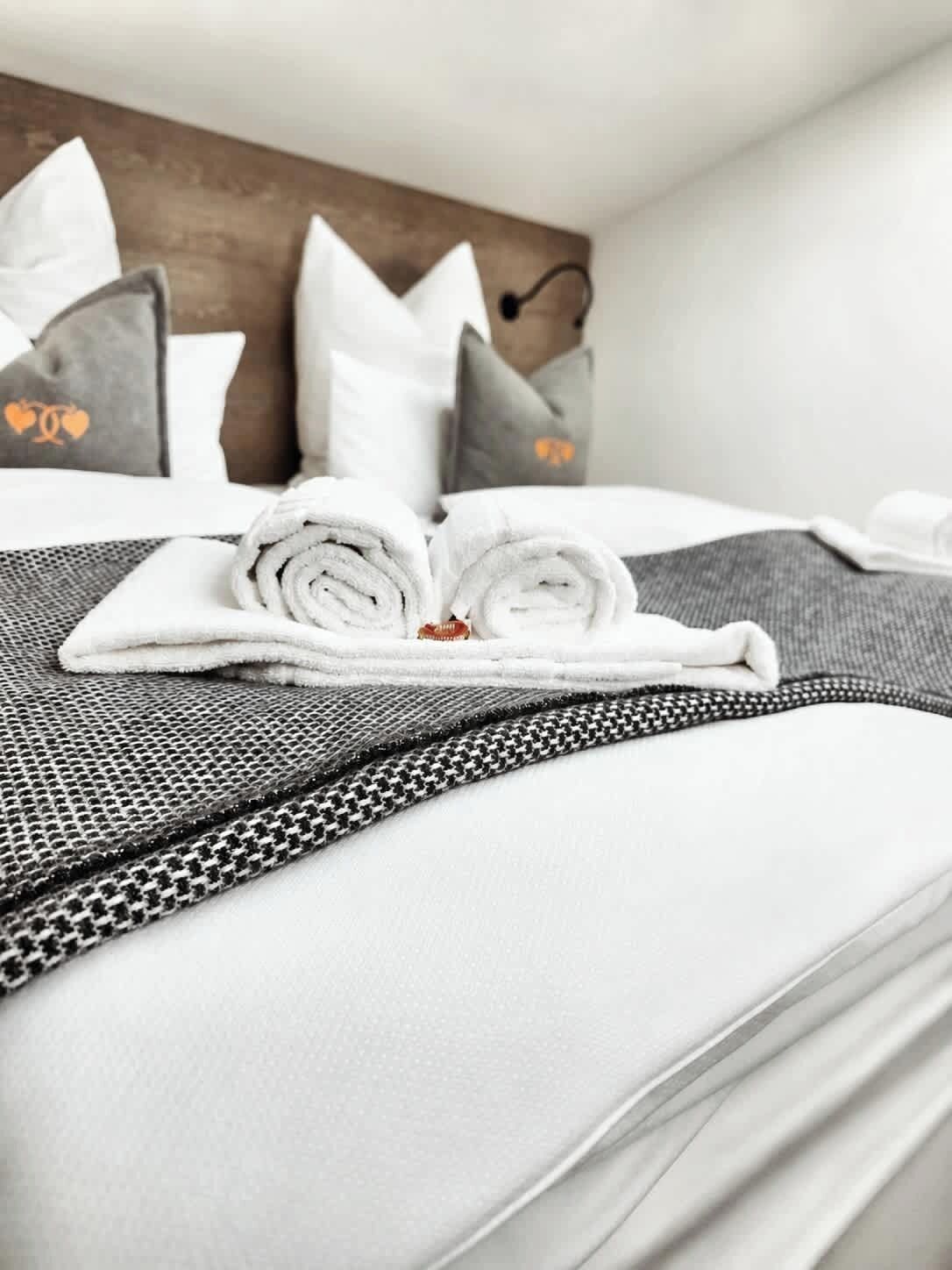 Een keurig opgemaakt bed met opgerolde witte handdoeken, een grijs beddegoed met patroon en passende kussens.