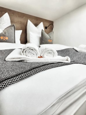 Un lit bien fait, avec des serviettes blanches pliées en rouleau, une couvre-lit grise à motifs et des coussins assortis.