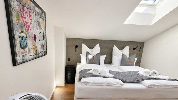 A bedroom with a bed, a standing fan, a skylight, and a wall art piece.