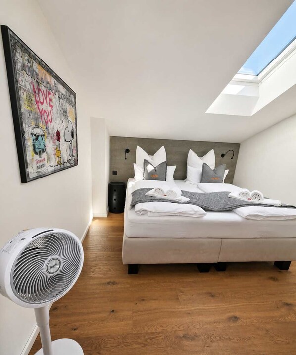 A bedroom with a bed, a standing fan, a skylight, and a wall art piece.