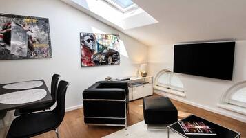 A modern loft bedroom with a skylight, a dining area with a bar stool, a black leather sofa, and a flat-screen TV mounted on the wall.