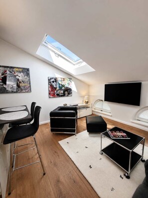 A modern loft bedroom with a skylight, a dining area with a bar stool, a black leather sofa, and a flat-screen TV mounted on the wall.