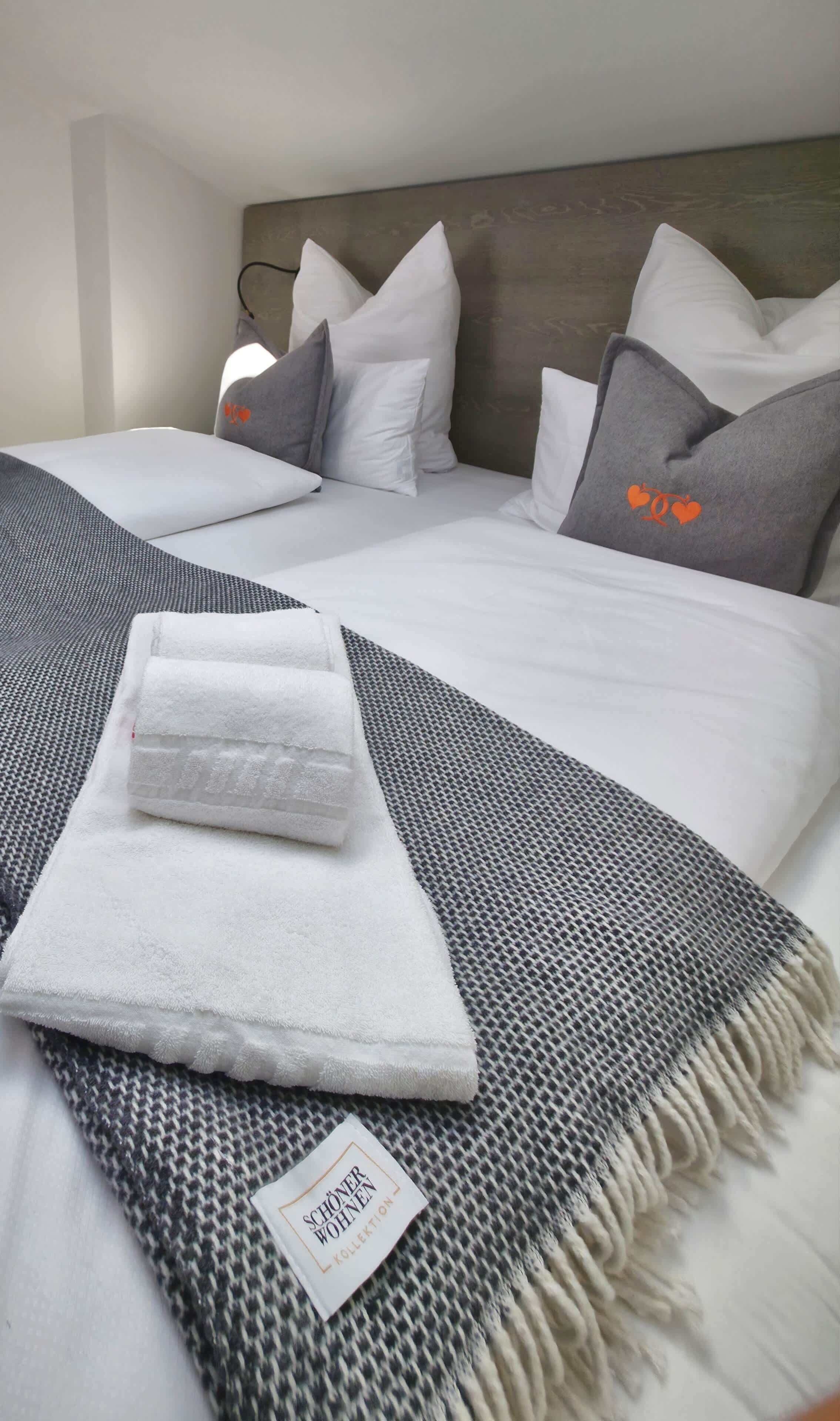 Una cama bien hecha con una manta a cuadros gris y blanca, almohadas blancas y una almohada gris con diseños de corazones naranja.
