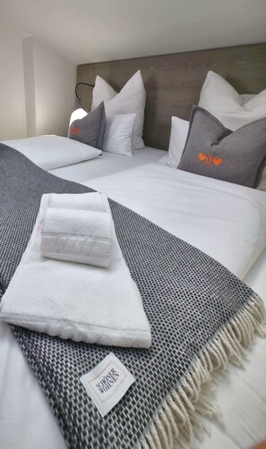 Cama bem arrumada com manta xadrez cinza e branca, almofadas brancas e almofada cinza com desenhos de corações laranja.
