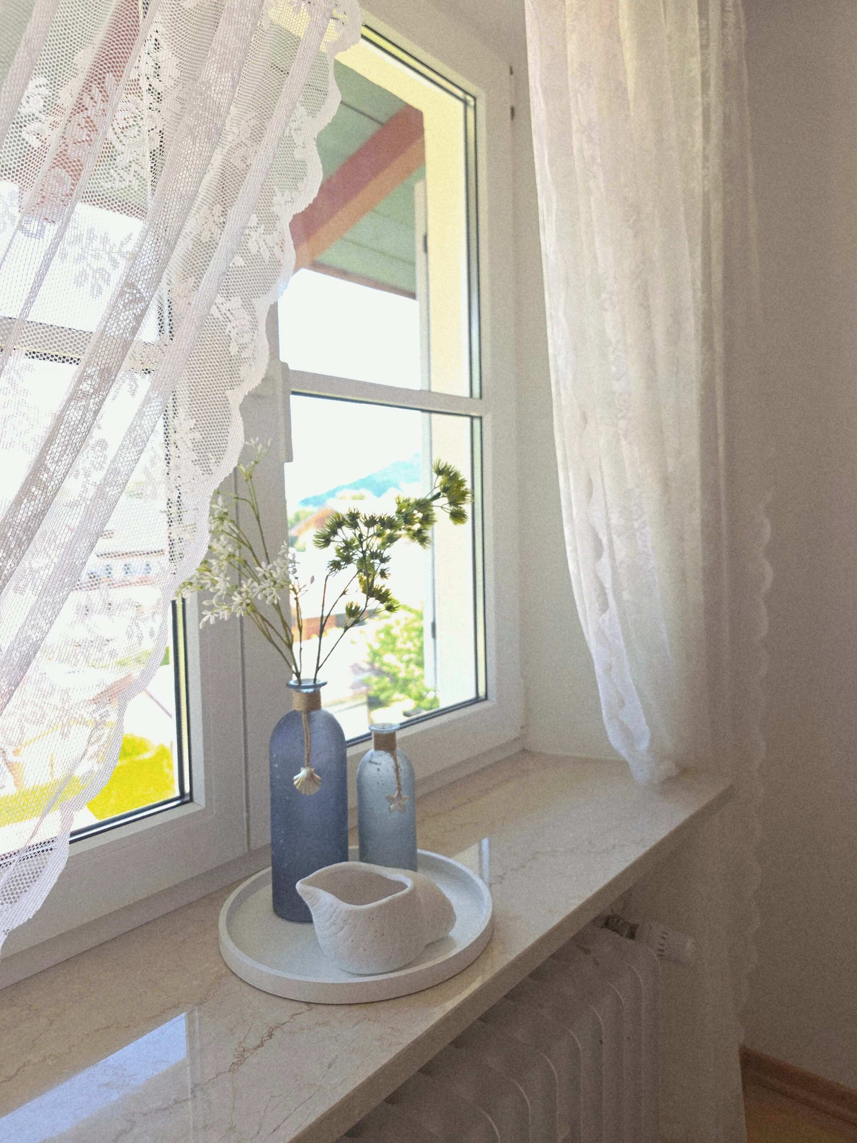 A window with lace curtains, two vases with flowers, and a small white dish on a marble ledge.
