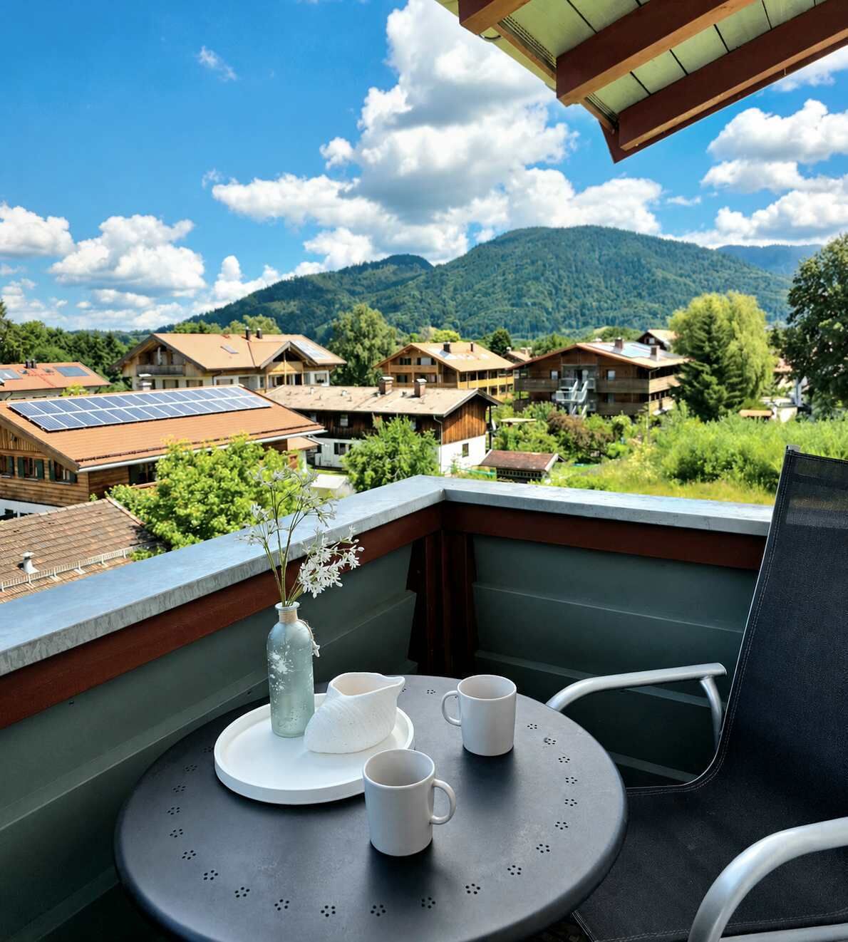 Een balkon met een tafel gedekt voor twee personen, een vaas met bloemen en een uitzicht op een dorp met huizen en bergen op de achtergrond.