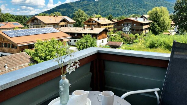 A balcony with a table set for two, a vase with flowers, and a view of a village with houses and mountains in the background. - 2 min to Tegernsee, Balcony, Sauna, Modern Kitchen (Bad Wiessee)