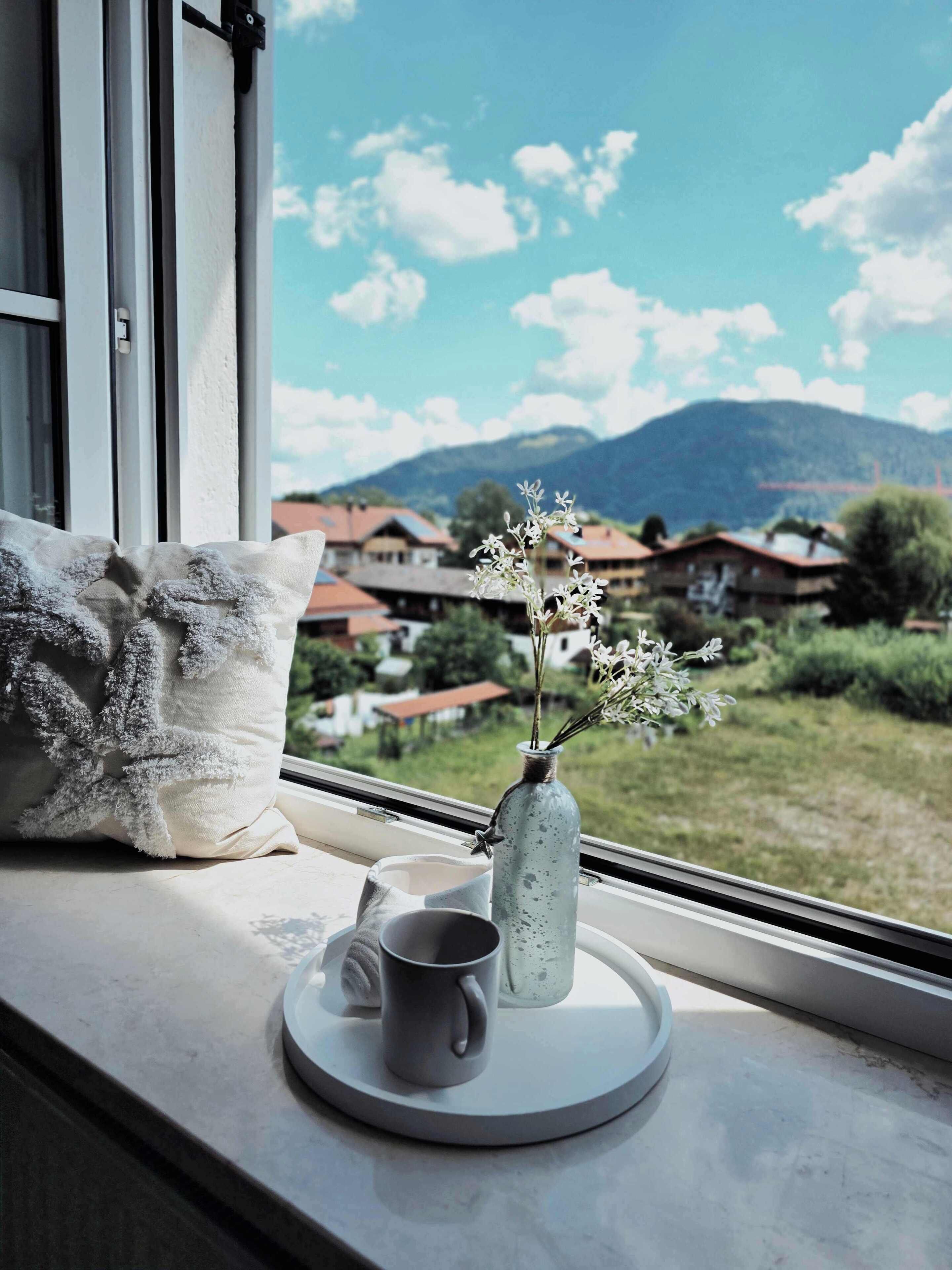 Ein Fenster mit Blick auf Häuser und Berge, ein Kissen, eine Tasse und eine Vase mit Blumen auf einer Ablagefläche.