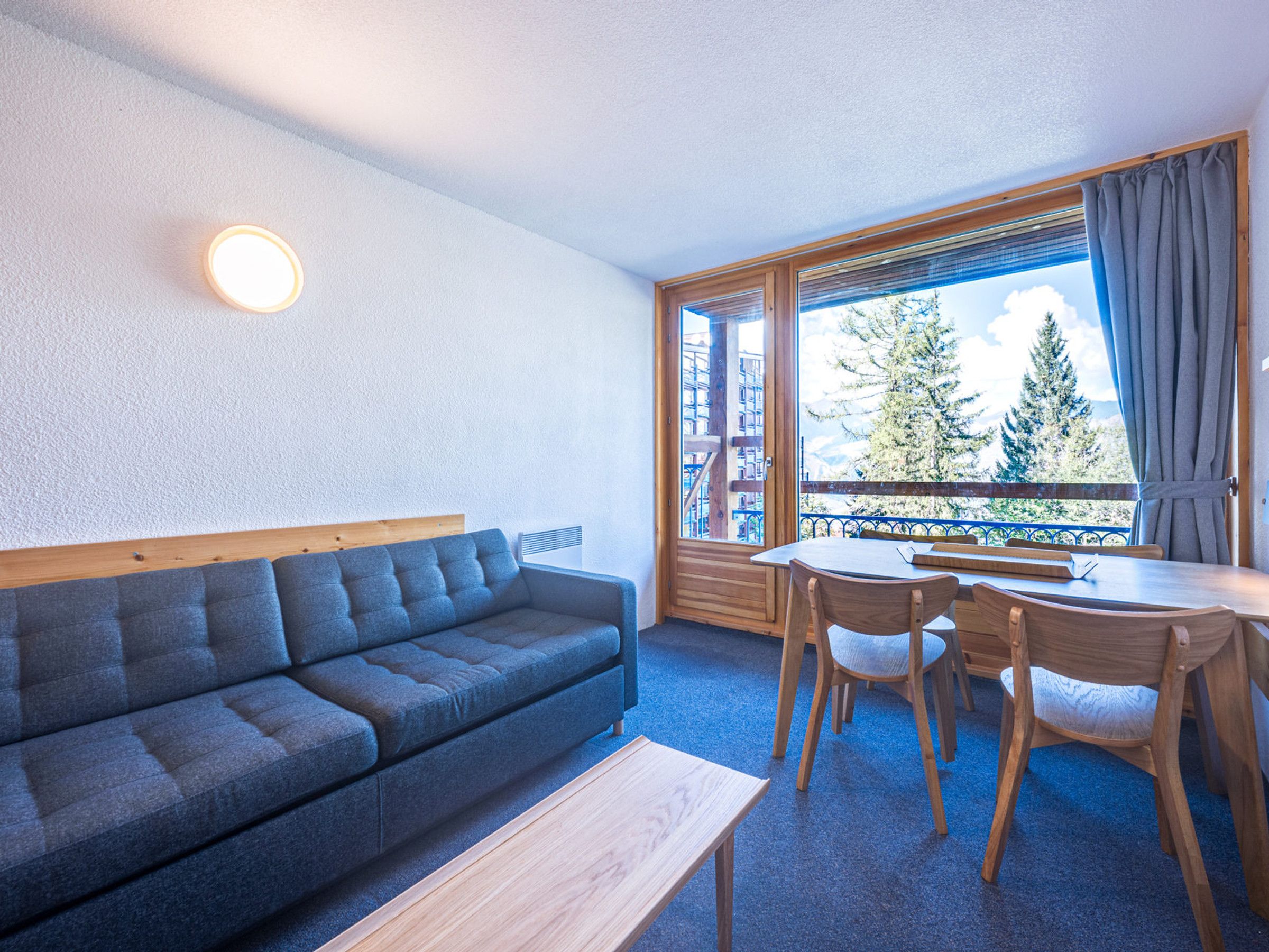 Ein modernes Zimmer mit einer Couch, einem Esstisch mit Stühlen, einem Fenster mit Vorhängen und Blick auf Bäume und Berge.