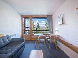 Een kamer met een bank, een tafel met stoelen, een raam met uitzicht op bergen en een wandklok.