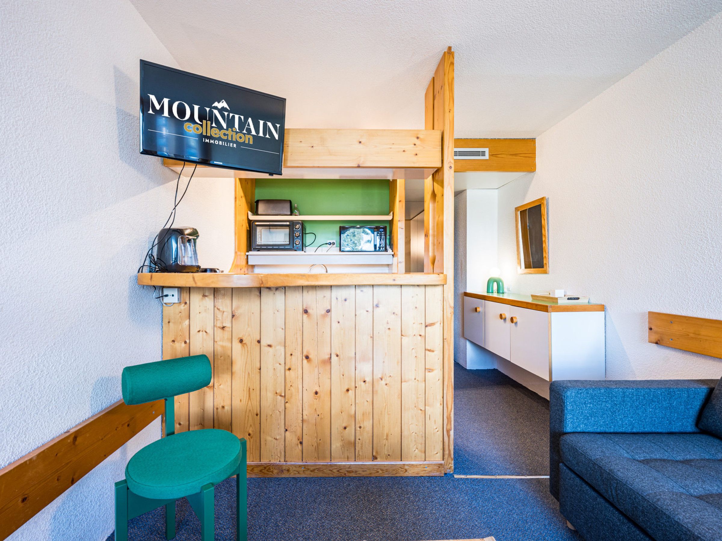 Una habitación compacta con una kitchenette de madera, un televisor montado encima y un sillón verde y un sofá azul.