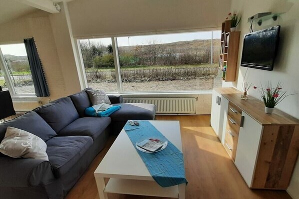 A modern living room with a sofa, a coffee table, a TV, and a view of a field through large windows. - Holiday home with 4 bedrooms (Julianadorp)