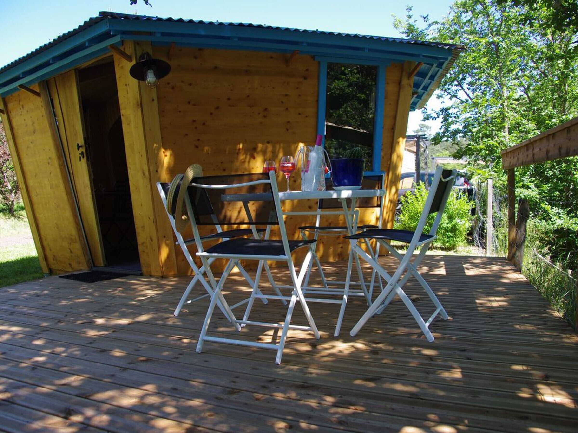 A wooden deck with a table and chairs, a covered area with a blue roof, and a wooden building with an open door.