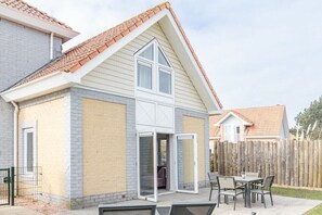 Un immeuble résidentiel à structure de deux étages, doté d’un patio couvert avec une table et des chaises, et d’une clôture en bois en arrière-plan.