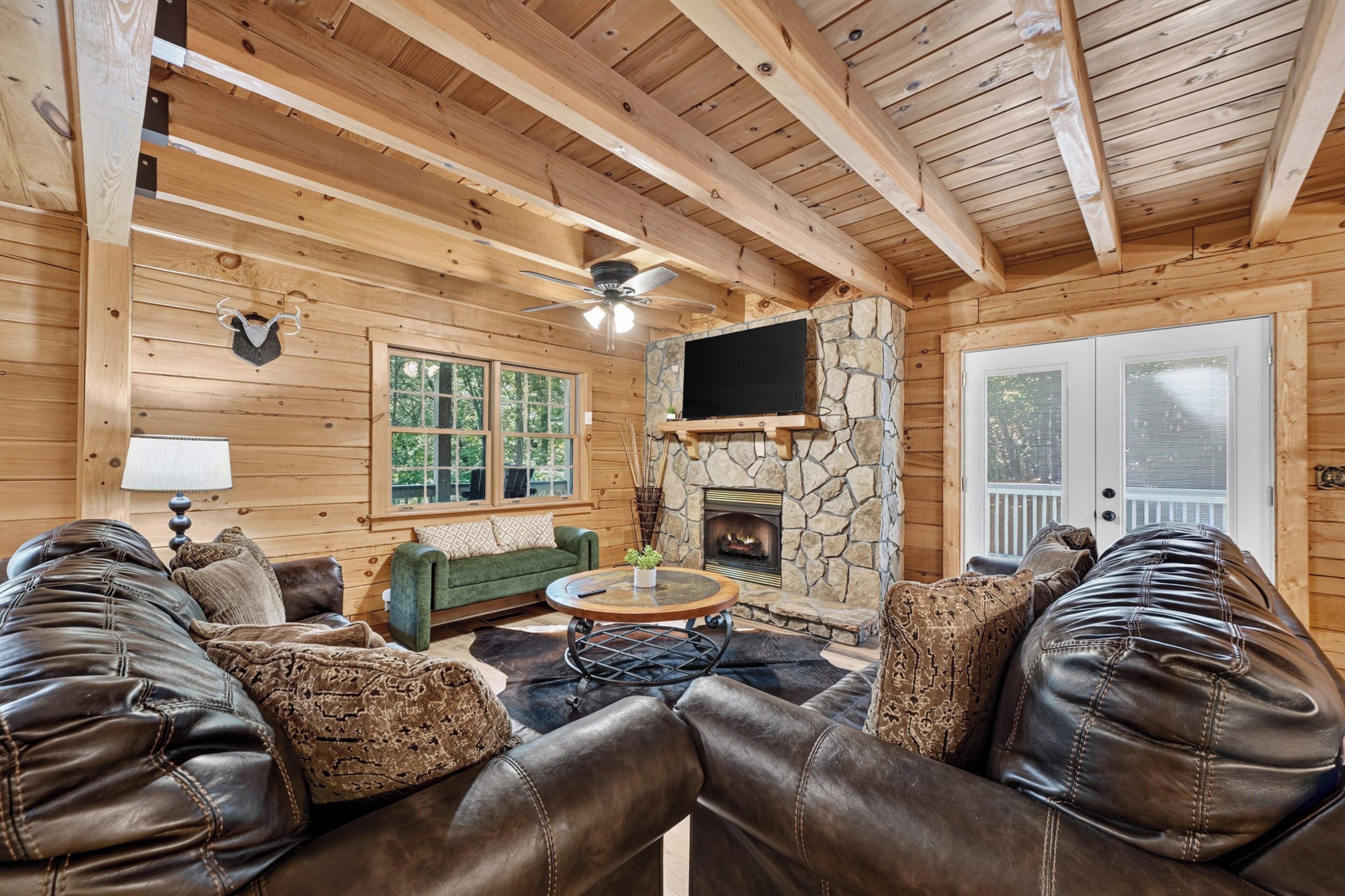 A log cabin interior with a stone fireplace, leather sofas, a television mounted above the fireplace, and a large window.