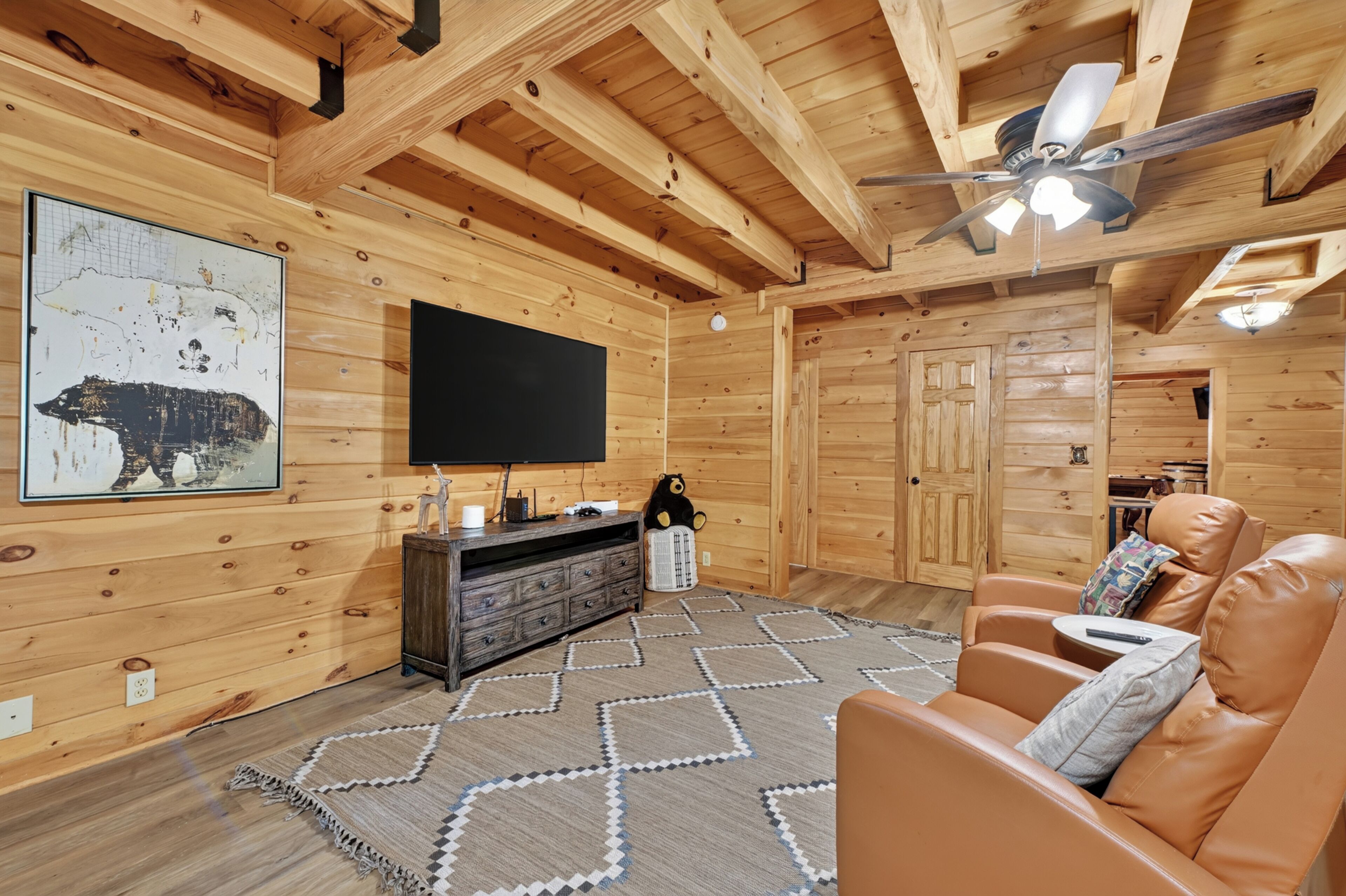A log cabin interior with a flat-screen TV mounted on the wall, a wooden entertainment unit, a patterned rug, leather seating, and a ceiling fan.