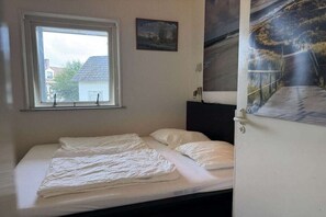 Ein Einzelbett mit weißer Matratze und Kissen, ein Fenster mit Blick auf Gebäude und eine Wand mit Landschaftsbild.