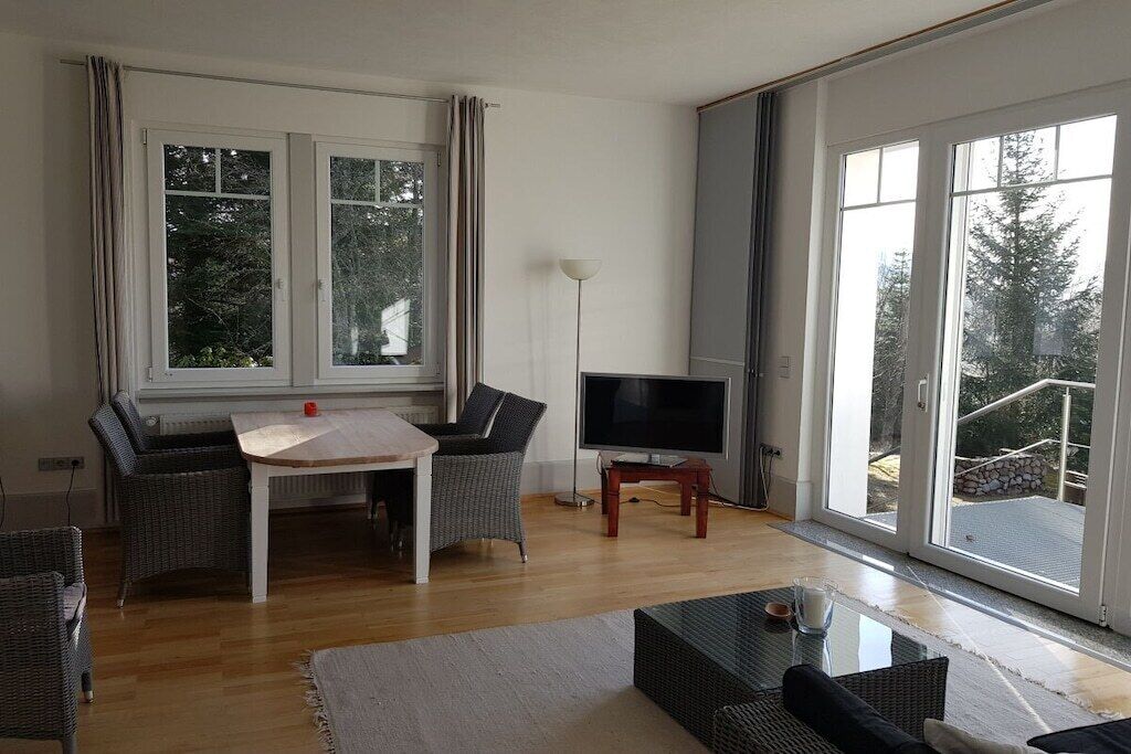 A living room with a wooden table, two chairs, a flat-screen TV, and a large window with a view of trees.