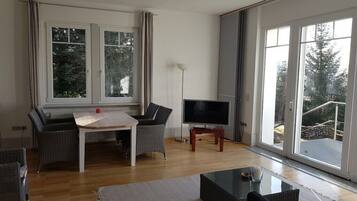 A living room with a wooden table, two chairs, a flat-screen TV, and a large window with a view of trees.