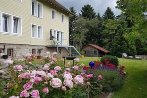 Ein zweistöckiges Haus mit einem Balkon, einem Garten mit rosa und roten Blumen und einem kleinen Schuppen im Hintergrund.