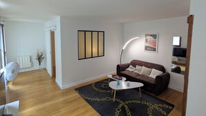 Un salon avec un canapé marron, une table basse blanche, un tapis à motifs noirs et jaunes, un ventilateur sur pied et une affiche accrochée au mur.