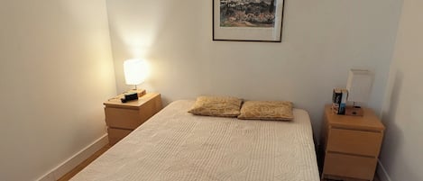 Quarto com cama, duas mesas de cabeceira, luminária na parede e um quadro emoldurado.
