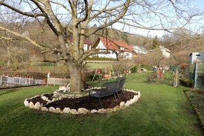 Ein Baum in einem Garten mit einer Bank, einem eingezäunten Bereich und einem Gebäude im Hintergrund.