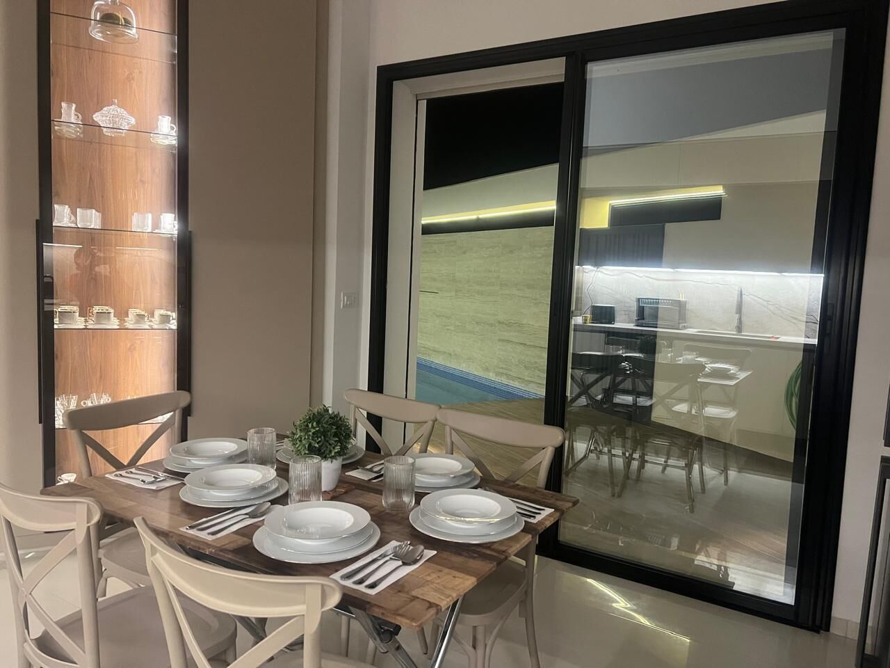 Un coin repas avec une table en bois dressée pour quatre personnes, des chaises, une plante en pot et une vue sur une piscine et un espace extérieur, le tout visible à travers une porte vitrée.