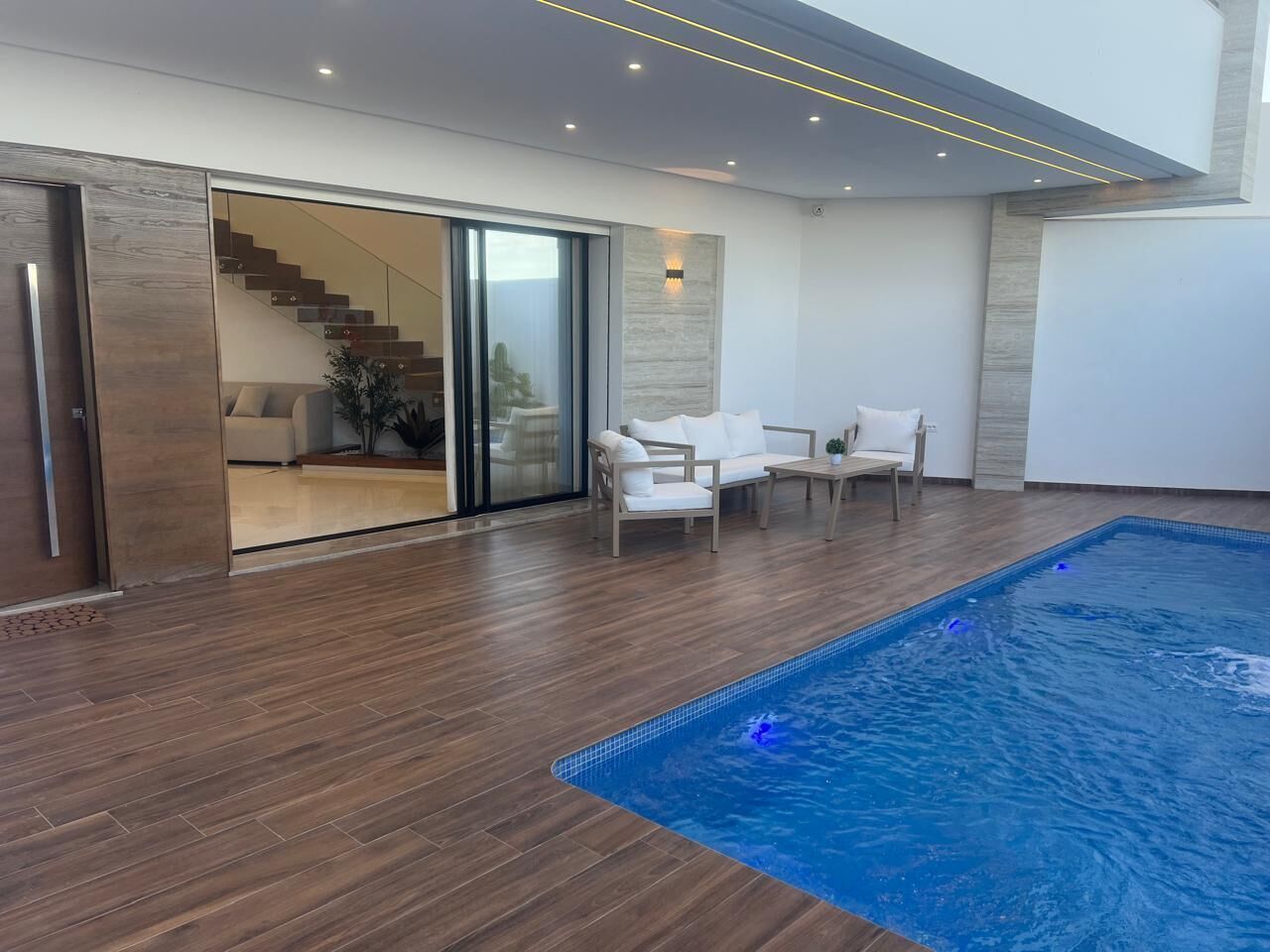 Une piscine intérieure moderne avec un plancher en bois, une porte vitrée donnant sur un escalier et un coin salon avec des chaises blanches.