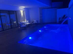 Un área de piscina interior moderna con fuente iluminada en azul, asientos y puerta de vidrio que da al exterior.