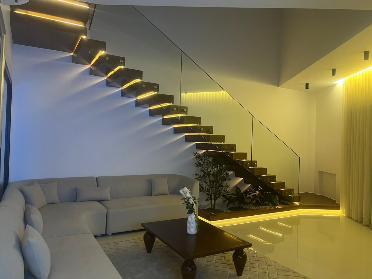 Un salon moderne avec un canapé aux lignes courbes, une table basse agrémentée d’un vase et un escalier doté d’un système d’éclairage intégré.