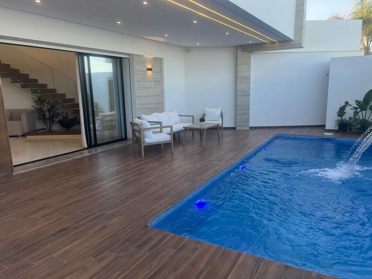 Un espace piscine intérieur moderne, doté d’un plancher en bois, d’un coin salon avec des chaises blanches et d’un escalier menant à un niveau supérieur.