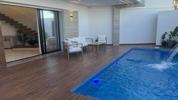 Un espace piscine intérieur moderne, doté d’un plancher en bois, d’un coin salon avec des chaises blanches et d’un escalier menant à un niveau supérieur.