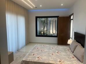 A bedroom with a large bed, a window with a view of greenery, and a door leading to another room. - VILLA ASIA (MAAMOURA)