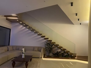 A modern living room with a curved sofa, a coffee table, and a staircase with integrated lighting. - VILLA ASIA (MAAMOURA)