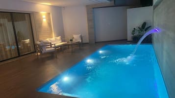 A modern indoor swimming pool with a waterfall feature, surrounded by a lounge area with seating.