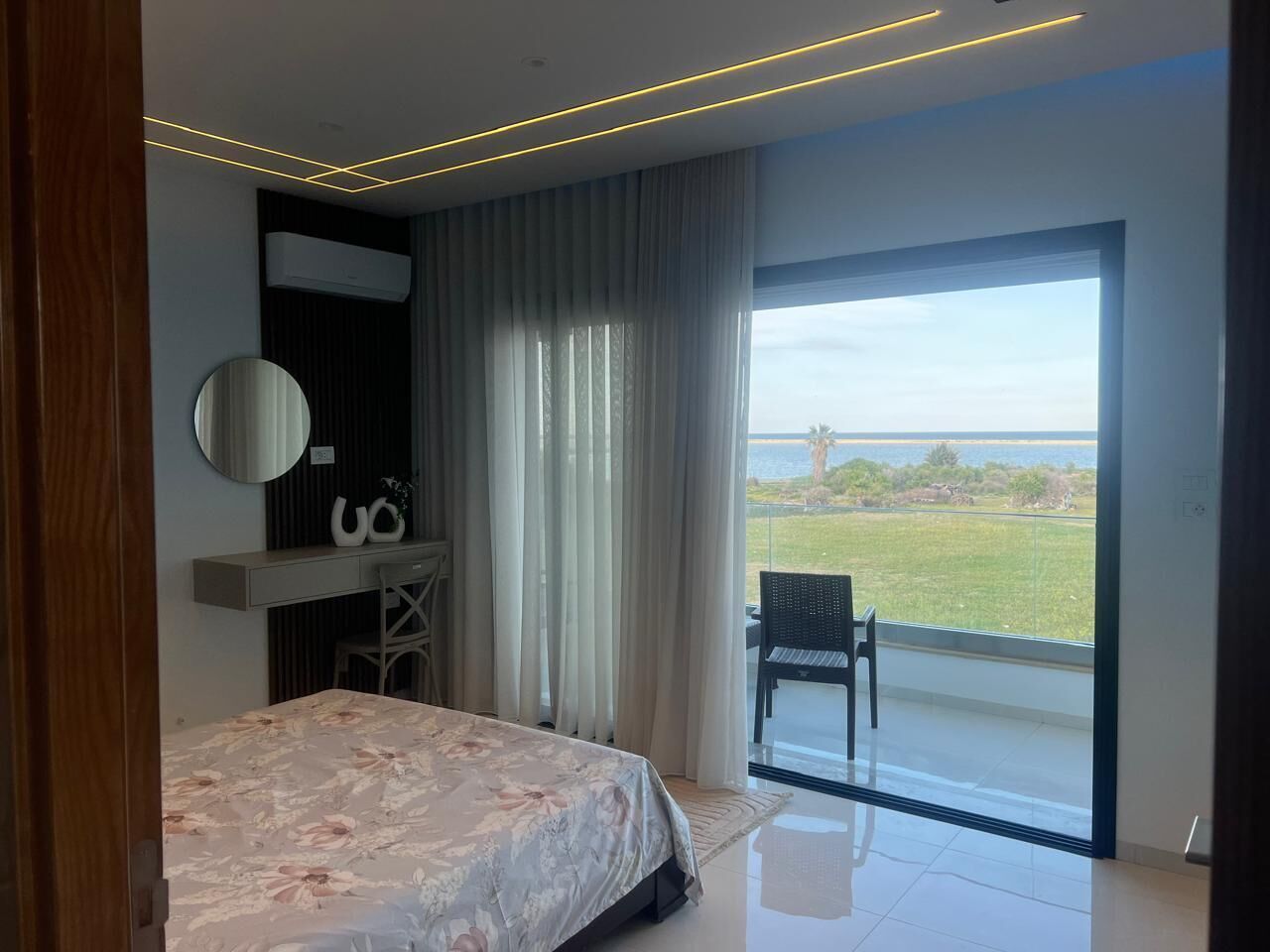 A bedroom with a bed, a desk, a chair, a mirror, and a view of the outdoors.